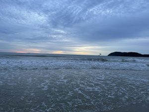 A peaceful evening view of the beach in Langkawi, Malaysia, with gentle waves washing onto the shore and soft sunset colors on the horizon. Distant islands and a parasail in the sky add depth to this serene coastal scene.